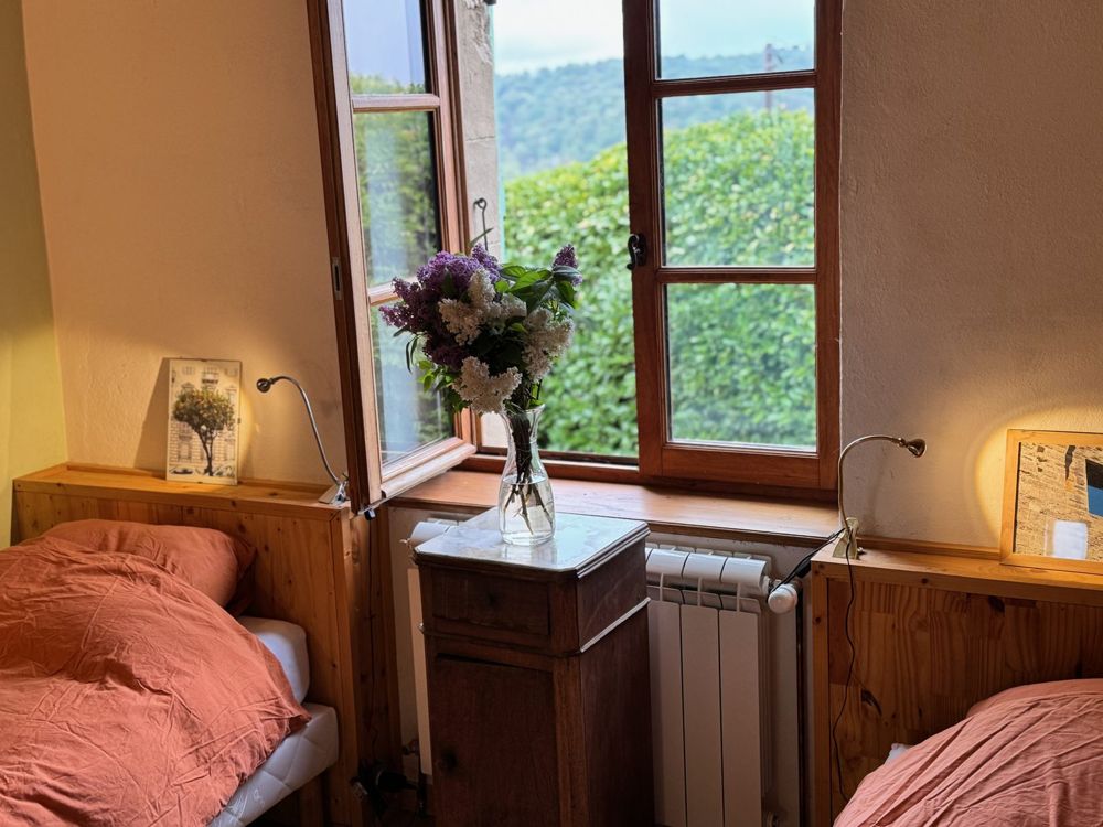 Chambre à deux lits séparés par une table de chevet fleurie de lilas, fenêtre ouverte sur la campagne