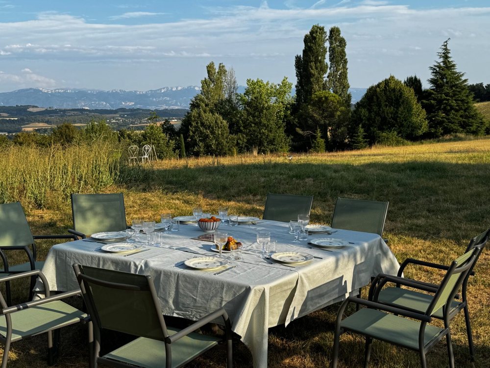 Table dressée avec nappe blanche et chaises vertes dans la prairie, le Vercors à l'horizon