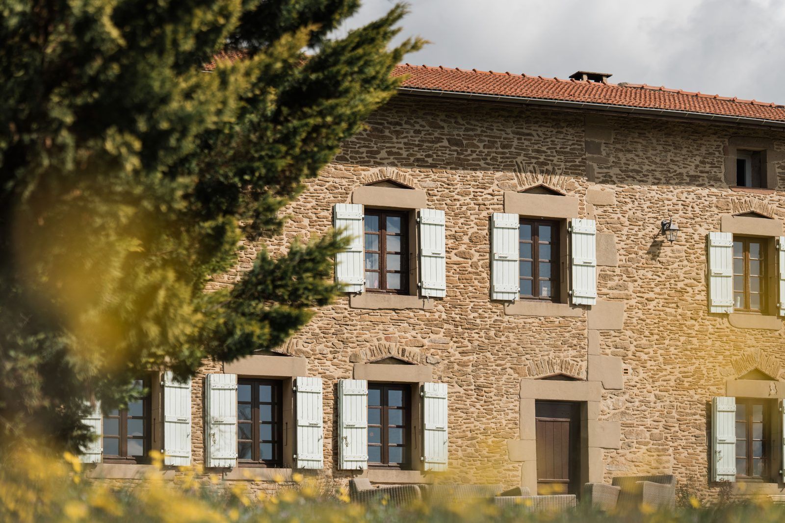 La façade en pierres dorées et ses volets clairs, derrière les herbes hautes