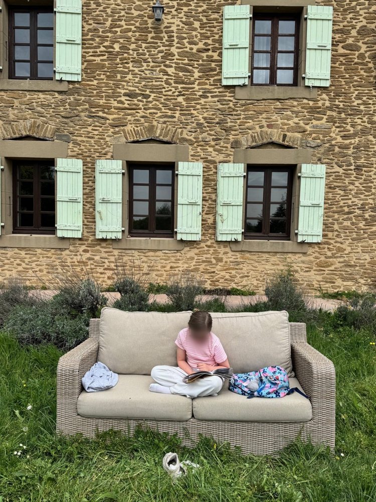 Une fillette lit sur le canapé de jardin devant la façade aux volets céladon